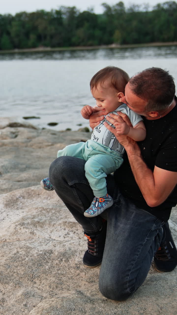 Father holding his little baby boy on one knee. Loving daddy kissing his son's cheek and back of the neck. Family concept. River at backdrop. Vertical video