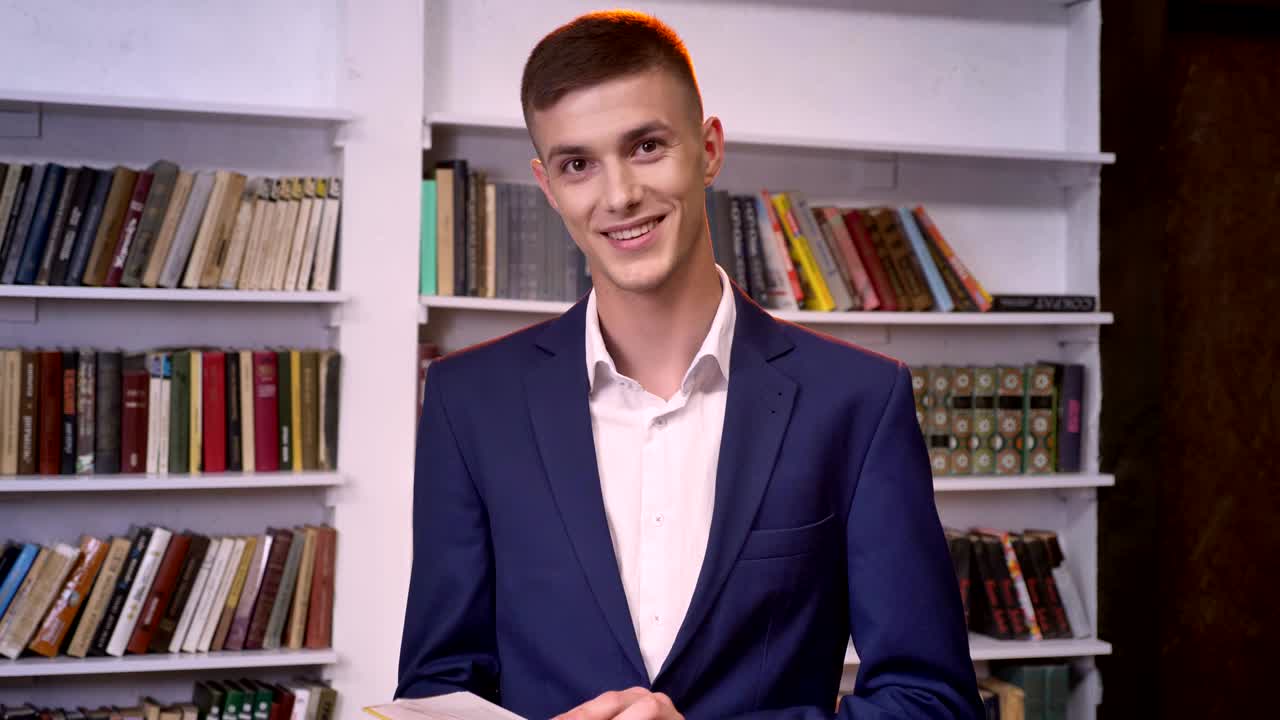 Young handsome businessman is reading book, watching at camera, smiling, bookshelf on background