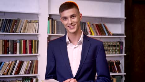 young handsome businessman is reading book, watching at camera, smiling, bookshelf on background
