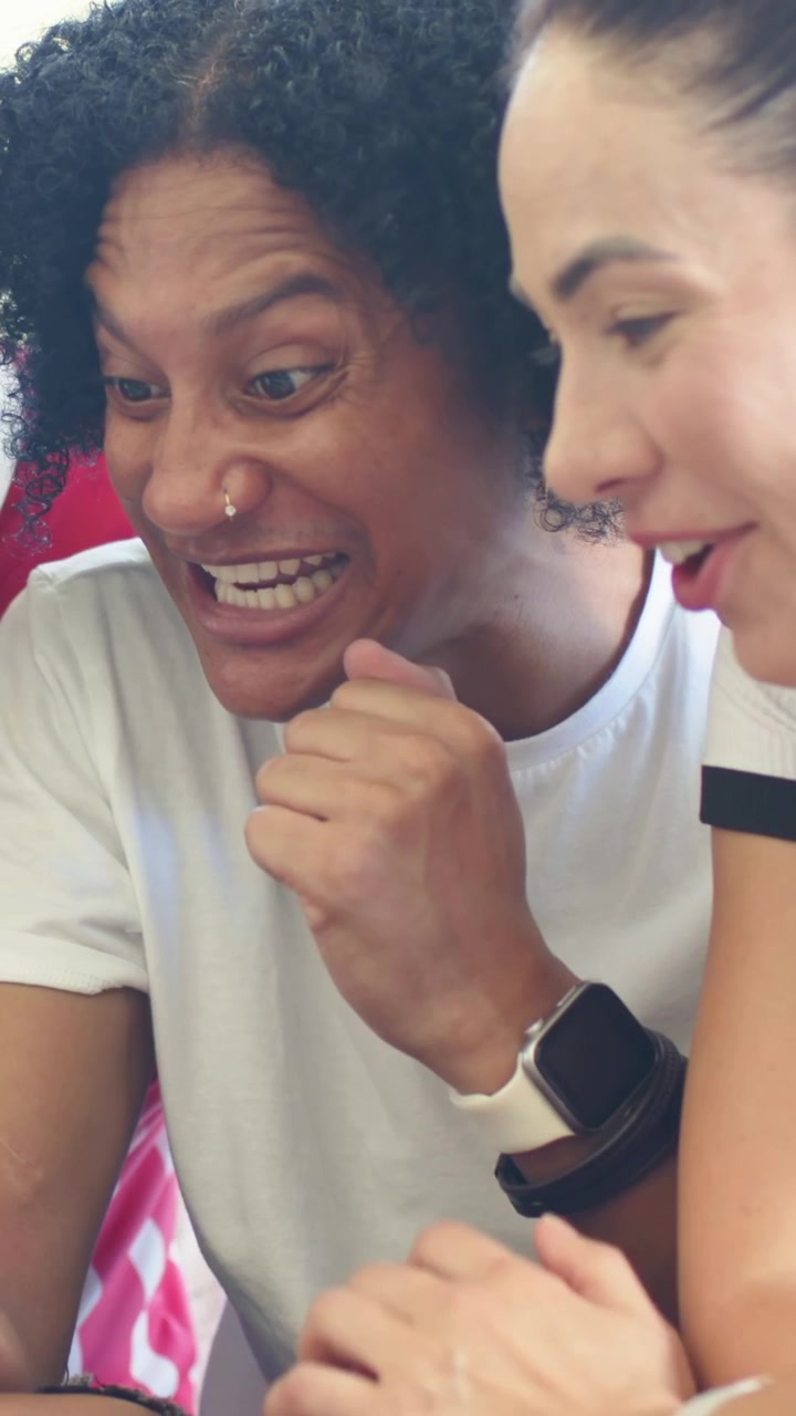 Vertical video: Reacting to phone lighting, two friends on red-sofa in white tee and polo, pointing