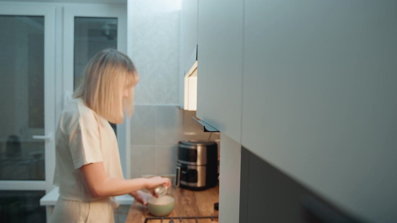 Blonde caucasian woman preparing breakfast in bright kitchen with daylight, mixing ingredients in bowl beside coffee maker, efficient focused pace, modern appliances, tidy counter, purposeful domestic