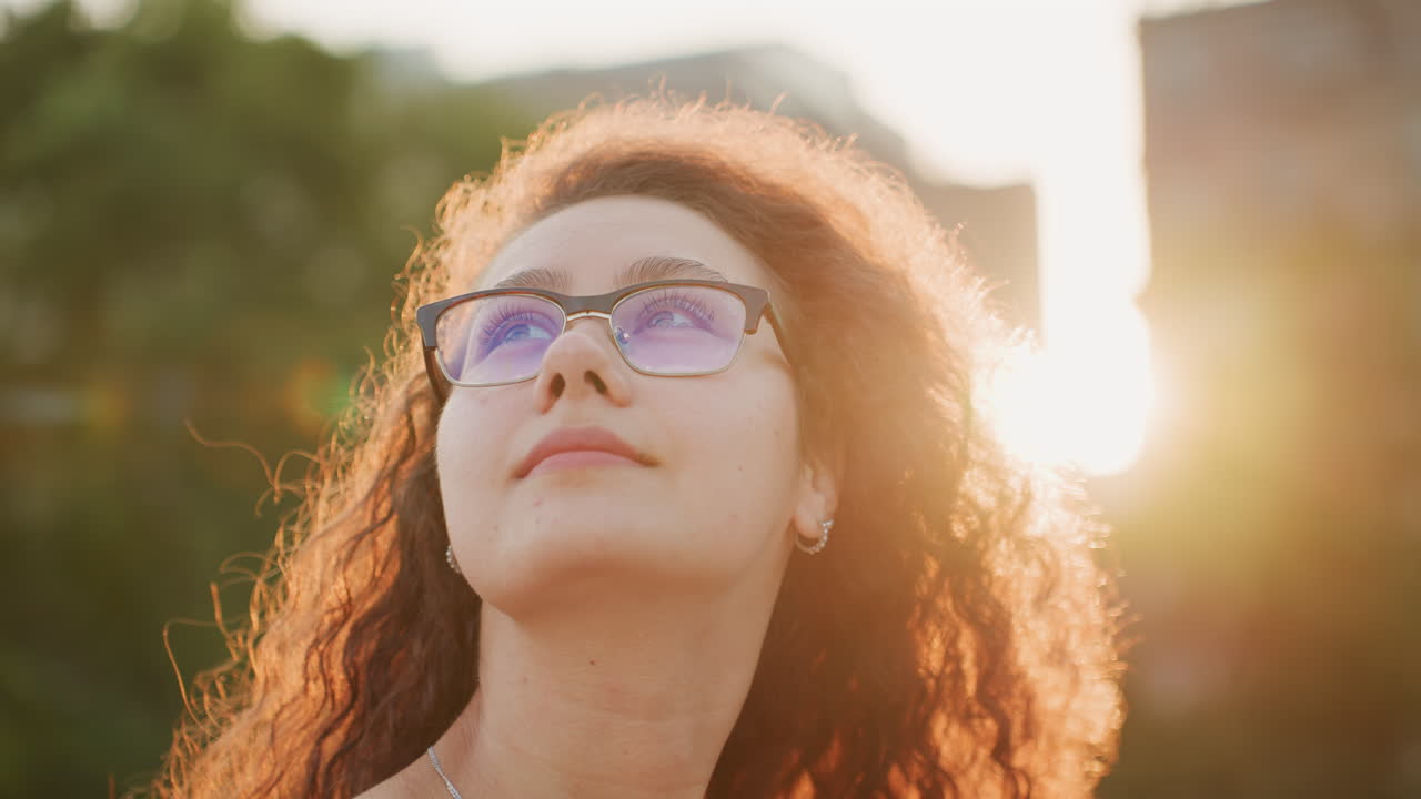 Mulher de óculos de perfil ao pôr do sol, halo de luz quente por trás ao redor do cabelo cacheado, horizonte da cidade borrado, olhar pensativo em direção ao horizonte, estudante desenhando no banco, pesquisadora fazendo uma pausa entre reuniões, momento suave.