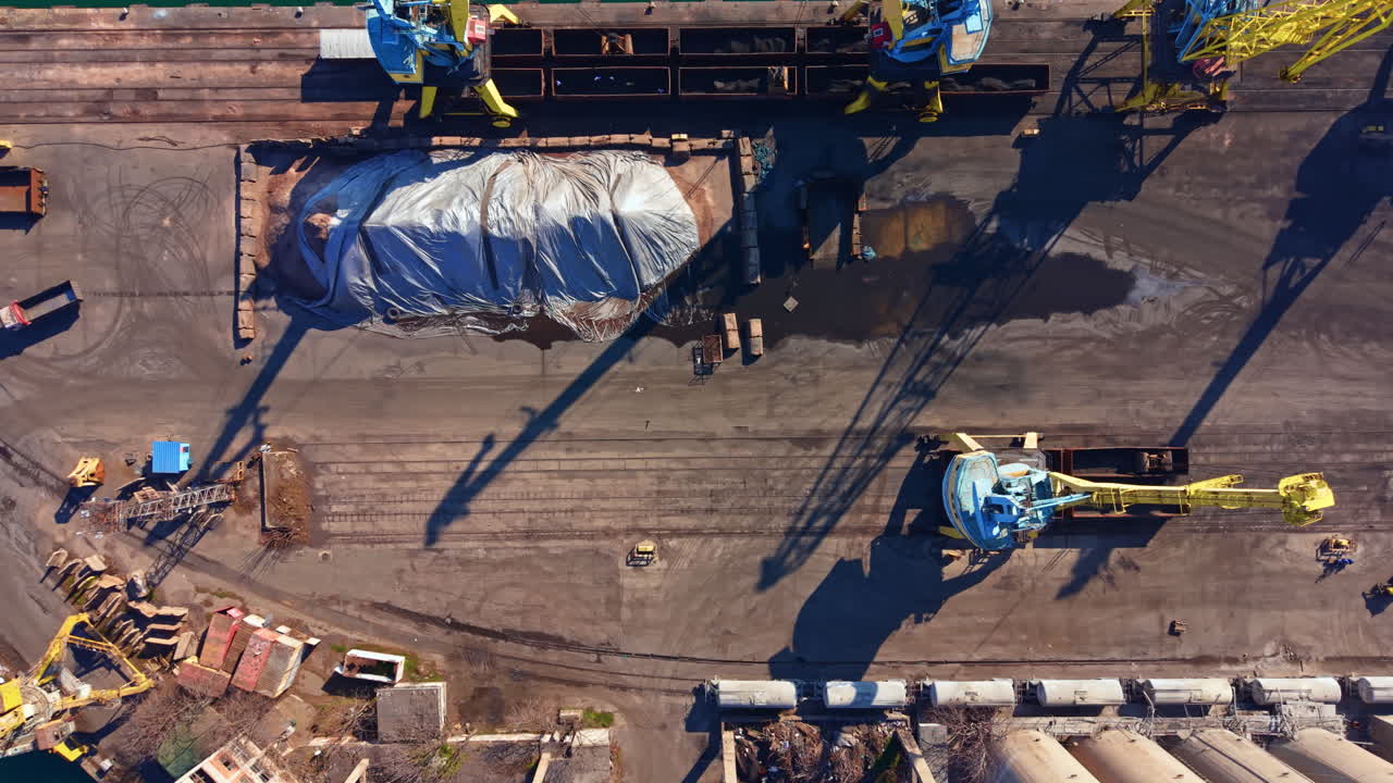 Cargo loading takes place at a busy port with large cranes working on stacks of materials in broad daylight over water