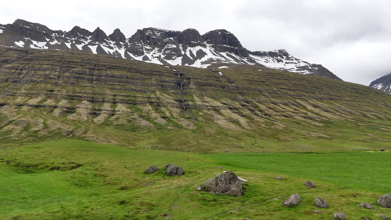 Wide aerial view of the mountains near Fáskrúðsfjörður, with snow-lined peaks, scattered waterfalls, winding wetlands, and an open valley crossed by a curving road under cloudy skies