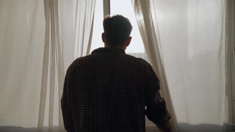 Man looking out window, back shot, peeking head between white curtains. Sun shining through curtains. Looking down at city street. Hiding and trying to avoid someone or something. Scared or curious