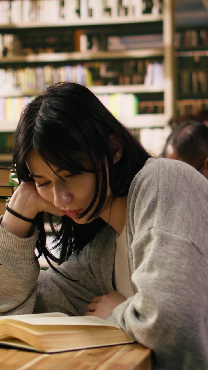 Video verticale: Studentessa stanca si appoggia alla mano mentre legge un libro aperto sotto una lampada verde