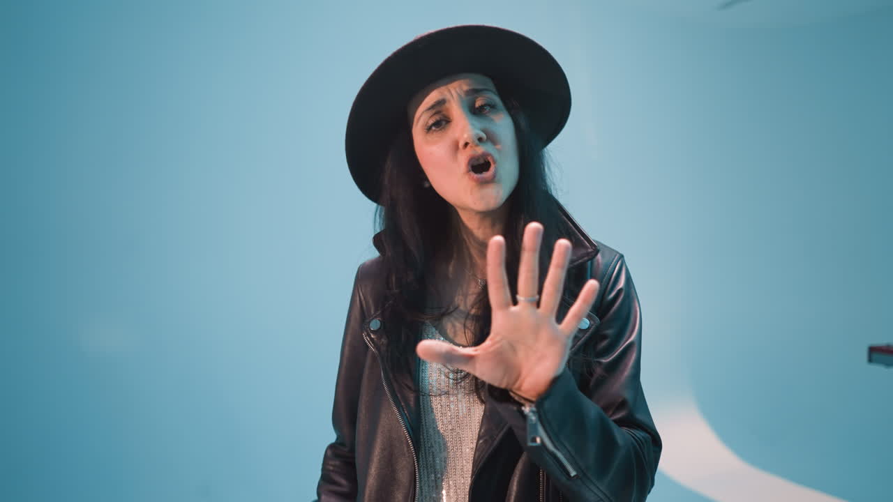 A woman in a black leather jacket, trousers, and hat sings passionately, moving her body, hands, and head. The camera zooms in slowly, against a blue background