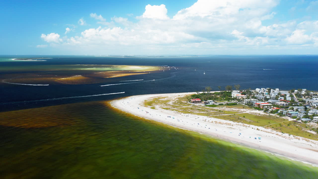 A sandy peninsula dotted with houses stretches along emerald shallows, where offshore sandbars break the darker sea, creating a vivid contrast of land and water