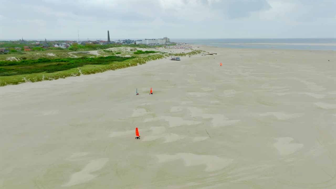 Colorful land yachts sail over vast sandy flats by Borkum’s shore under overcast skies. Aerial orbit view
