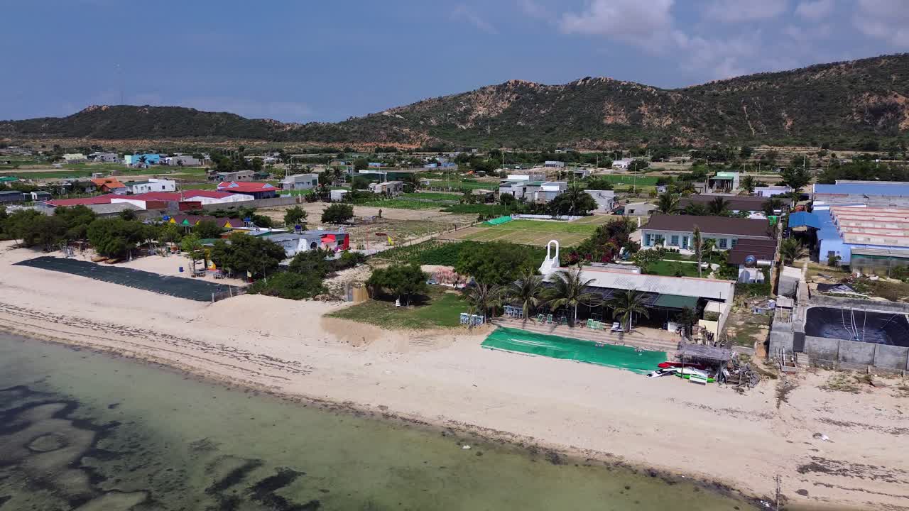 Dronie of a beach resort in Phan Rang, Vietnam, showing white sand, a pool, and revealing the horizon on a sunny day.