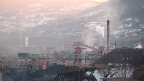 the morning haze lingers over an industrial site with smokestacks and a large coal pile, set against a backdrop of snow-dusted mountains