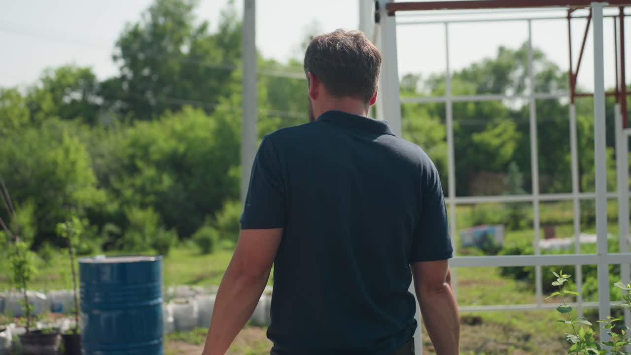 side view of man in blue polo carrying wooden crate and work tool walking past white metal frame near green garden plants on sunny day inspecting outdoor project