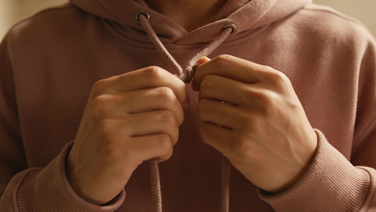 Mani adulte che afferrano i cordoncini, stringendo un nodo per fissare i lacci di una felpa rosa antico a casa.