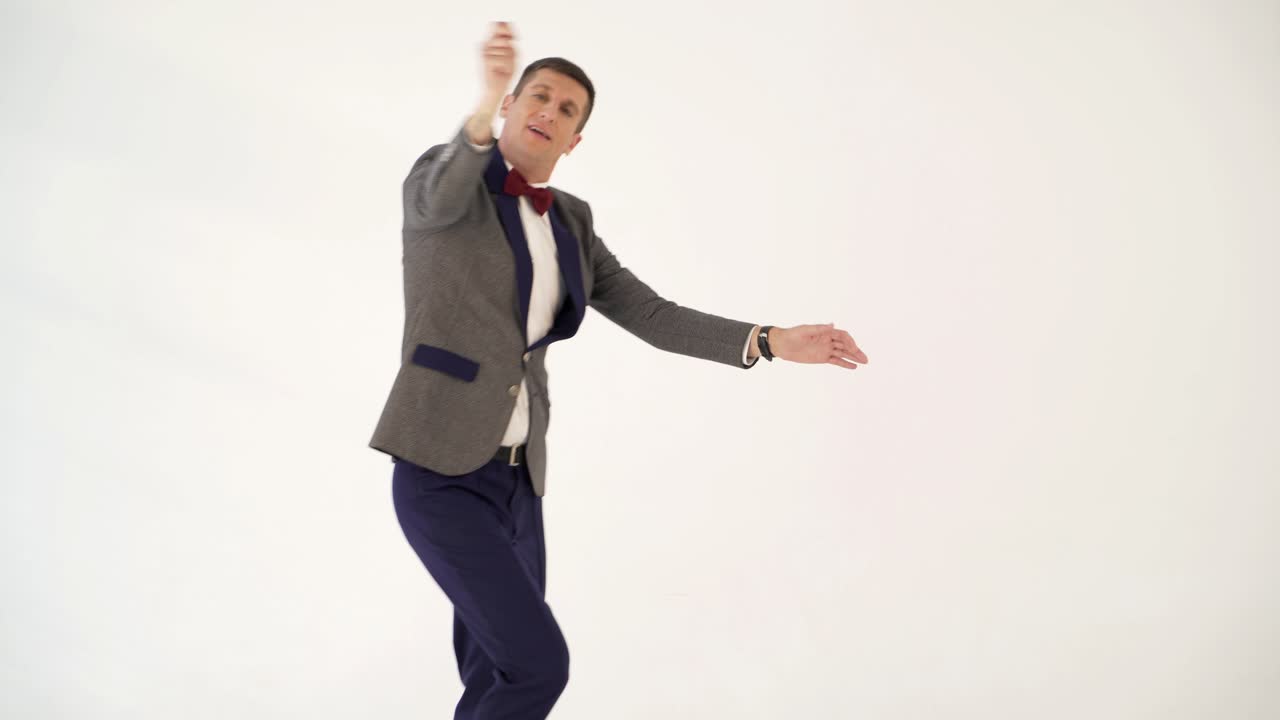 humorist in gray suit with red bow tie is dancing in the similitude of dancer with ribbon in hand on white background in studio