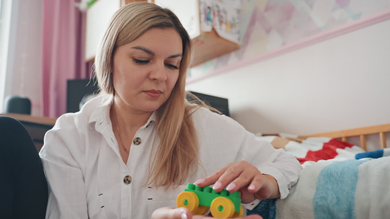 Mamma caucasica sistema un giocattolo per il figlio in una accogliente cameretta, testando un'auto di legno colorata con espressione concentrata e delicata, indossando una fascia per capelli e una camicetta bianca, seduta sul letto con coperta e scaffali dietro, atmosfera calda.
