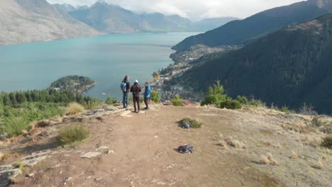 slowmo - tres jóvenes viajeros parados en la caminata por la colina de queenstown, nueva zelanda y mirando el lago wakatipu, montañas con nieve fresca y nubes - drone aéreo