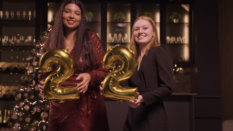 two women friends wearing elegant clothes holding balloons with the numbers 22 while dancing in new year's party 1