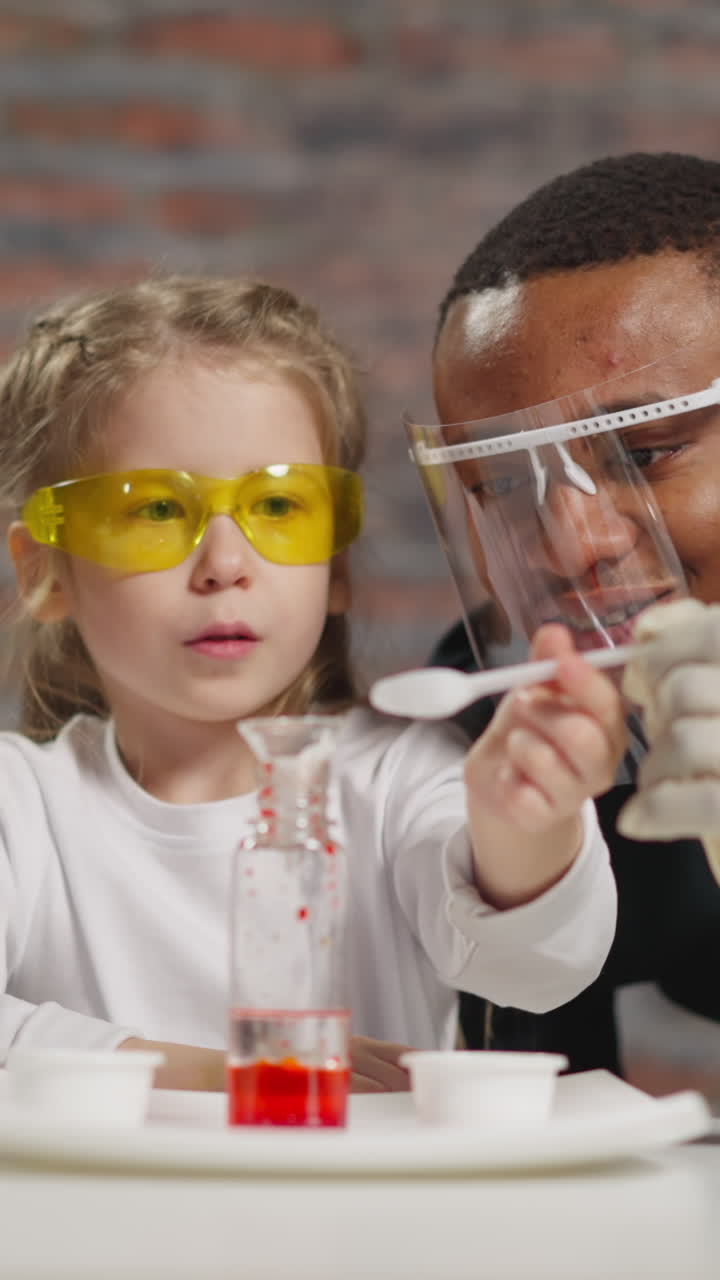 Little girl student adjusts funnel on bottle with liquid and stirs substance doing funny tests with Asian and African-American teachers in lab