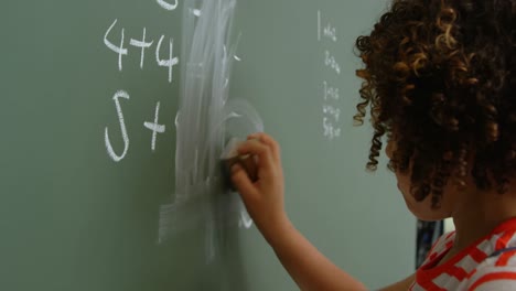 Side-view-of-mixed-race-schoolgirl-cleaning-green-chalkboard-in-classroom-at-school-4k