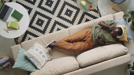 Afro-American-Man-in-Headphones-Using-Smartphone-on-Sofa