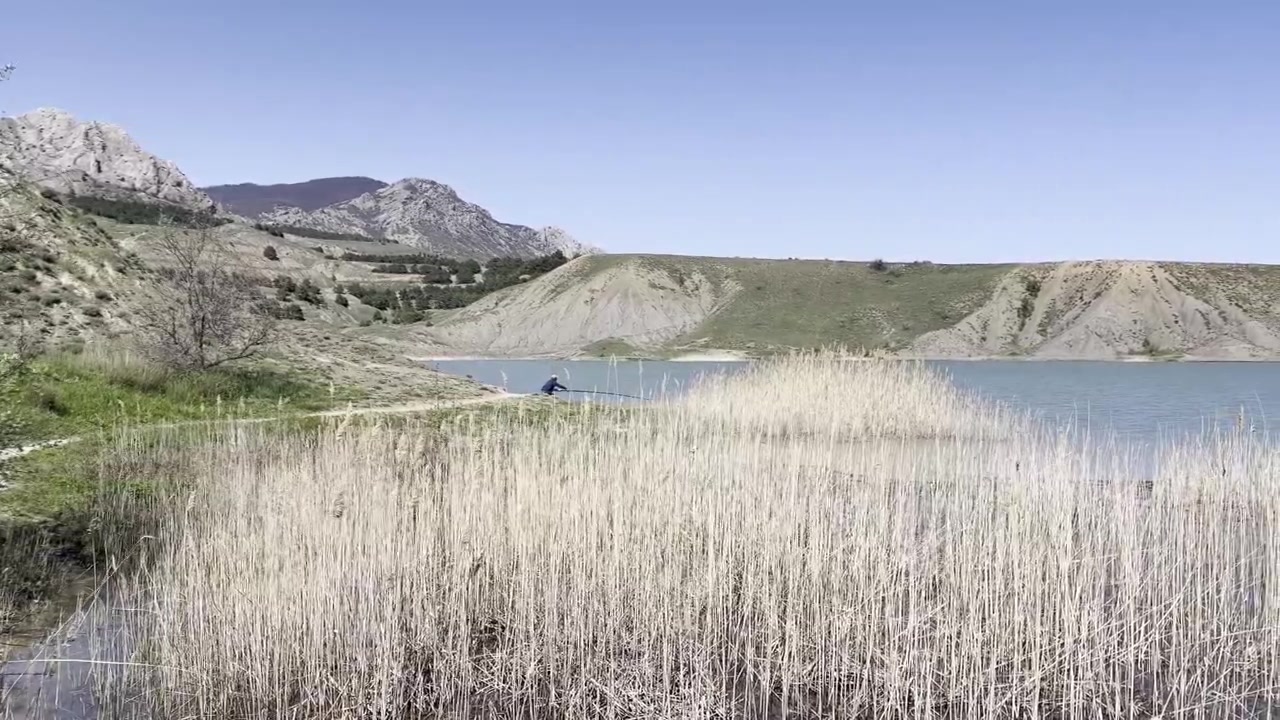 Un pescatore solitario getta la sua lenza in un lago calmo, circondato da canne secche, verdi colline e montagne lontane sotto un cielo azzurro e limpido.