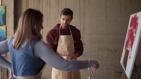 female artist teaching young male student to paint with oil paints. dark haired man putting on beige apron, getting ready for lesson. workshop at studio