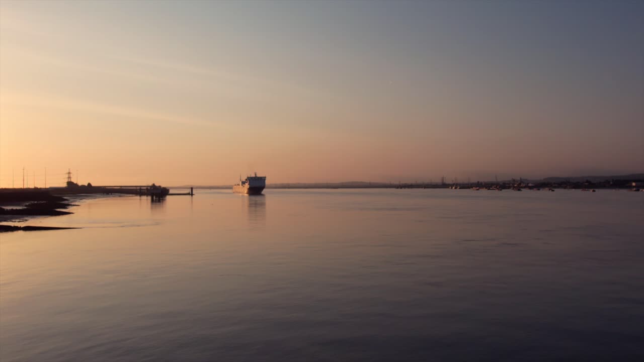 A cargo ship moves along the Thames River at sunrise, reflecting the warm light on calm water. Industrial structures and power lines are visible in the background. Droneshot