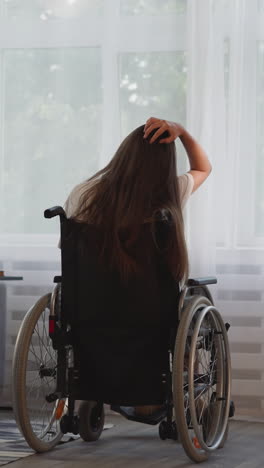 una mujer solitaria con discapacidad con esclerosis múltiple mira por una ventana brillante en la habitación. una mujer morena se sienta en una silla de ruedas tocando el cabello largo y suelto vista trasera