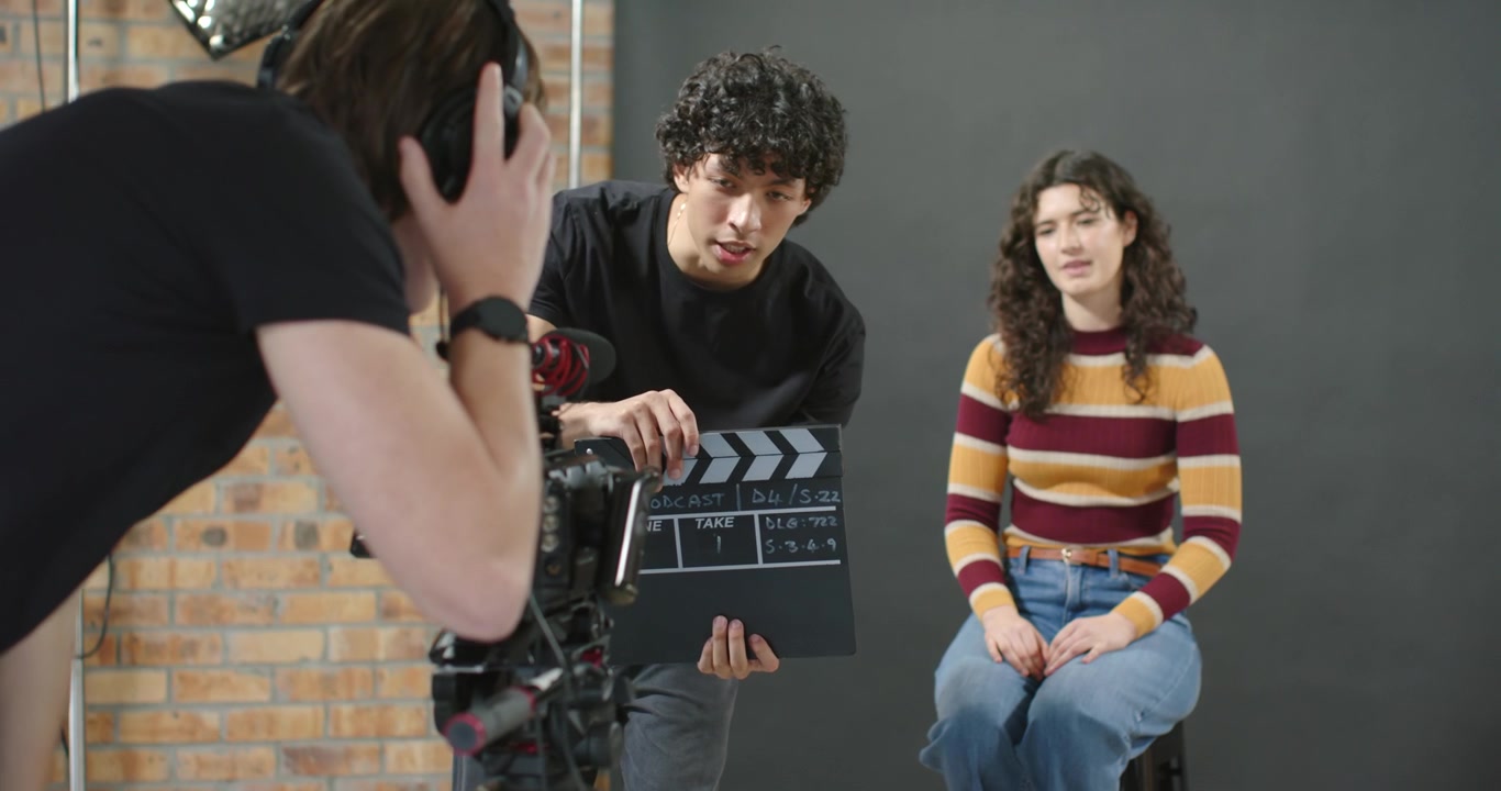 Troupe cinematografica che registra una scena, il ciak che si chiude, la telecamera inquadra una donna con un maglione a righe seduta su uno sgabello