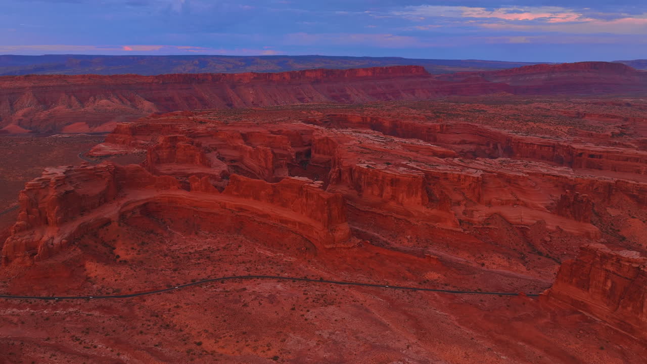 Approaching the stunning red huge canyons in the vast desert. Cars ride by the highway around the rocks. The Arches National Park, Utah, USA at sunset