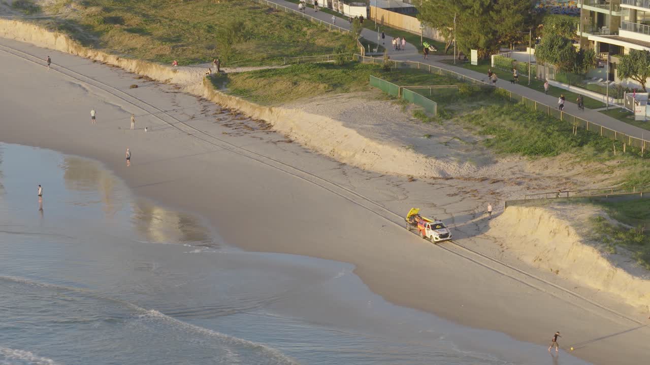 Aerial view of a patrol vehicle driving along Gold Coast beach with people walking nearby, under soft daylight