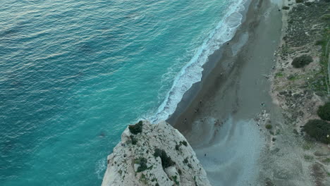 Aerial-view-of-a-coastal-landscape-at-dusk,-showcasing-a-winding-road-beside-a-serene-beach-with-gentle-waves-and-a-steep-cliff-edge-overlooking-the-sea