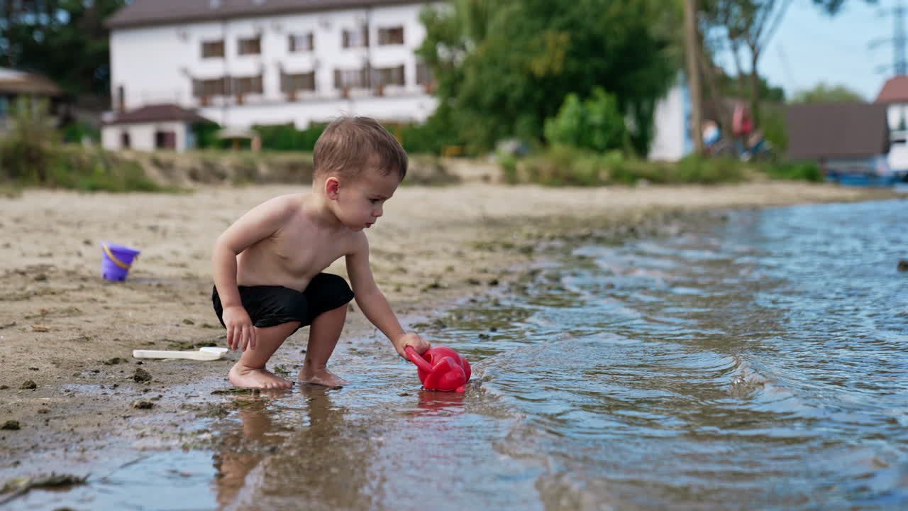 Caucasian toddler wearing black shorts filling the watering can. Baby boy playing near the river in summer.