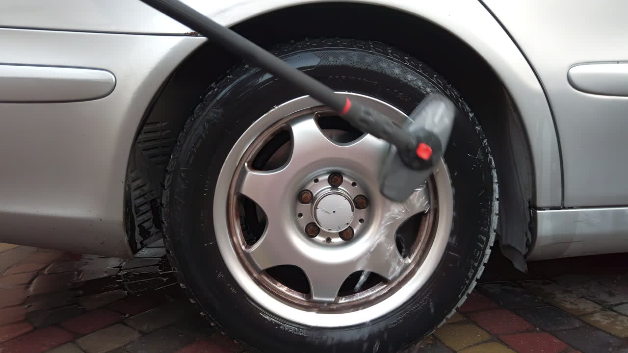 A person uses a pressure washer to clean a car tire outside. The shining rim and wet surface show the dirt being removed effectively. This activity happens in a bright, sunny environment