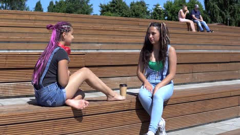 two girl friends talking to each other having a good mood sitting on a bench in the park