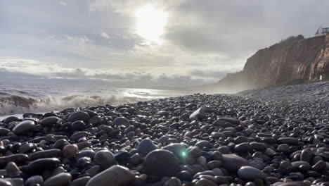 Shot-of-a-pebble-beach-with-the-waves-crashing-on-the-coast-line,-shot-with-a-setting-sun-in-the-sky