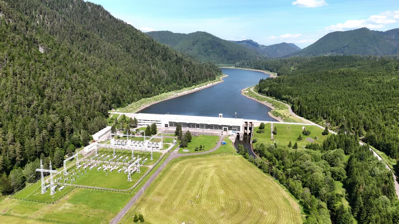 Drone approaching and descending toward Čierny Váh dam, revealing detailed view of the hydroelectric plant, blue reservoir, and surrounding green mountain landscape in Slovakia