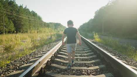 back view of little boy in dark shirt and shorts walking along rail track surrounded by gravel, grass, and forest under clear sky with warm sunlight casting soft glow over peaceful countryside