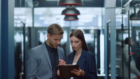 Young-colleagues-discussing-project-in-corridor.-Man-and-woman-looking-at-charts