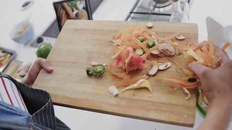 midsection of biracial woman in apron composting vegetable waste in kitchen, slow motion