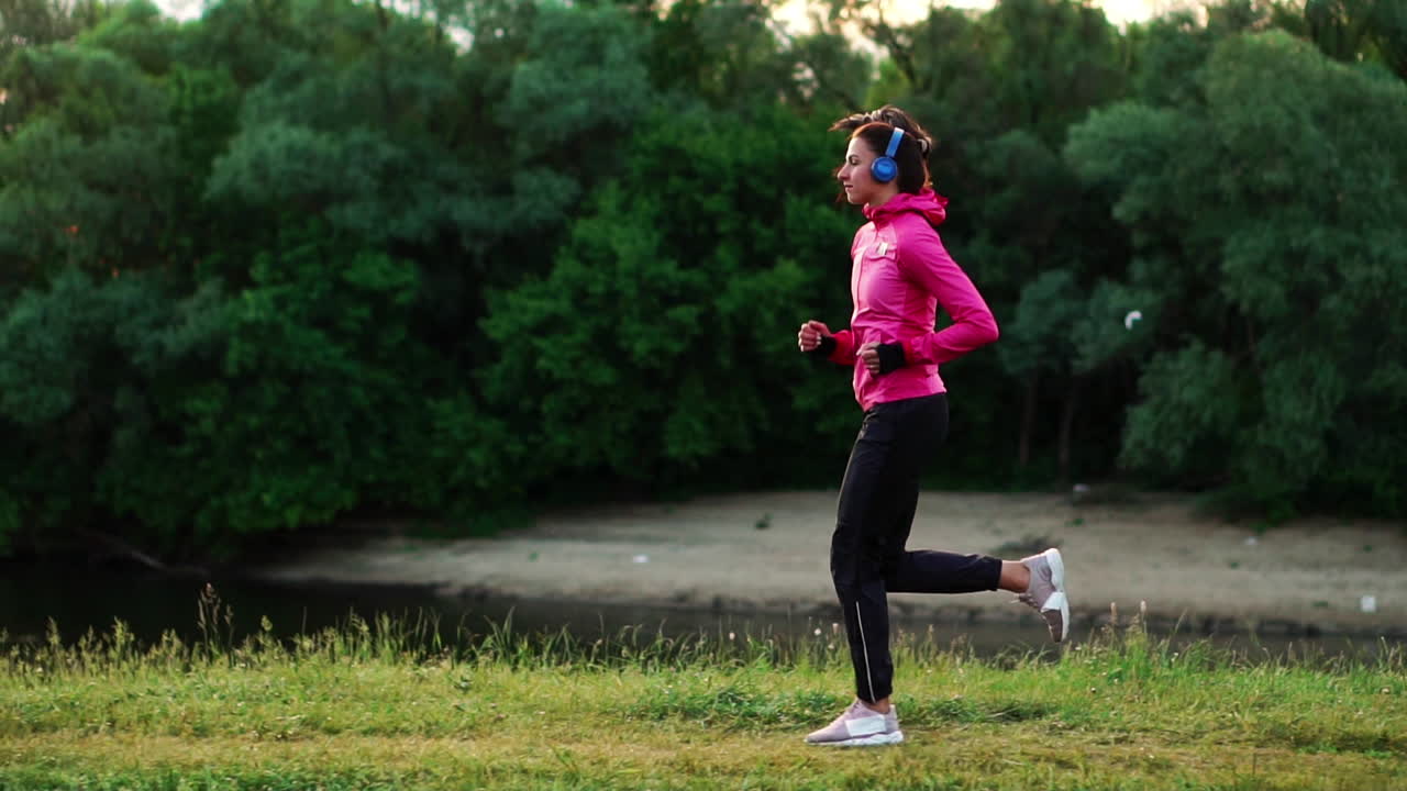 Brunette with long hair in headphones runs along the river in the Park in the morning at sunrise in the summer in a pink jacket and black pants