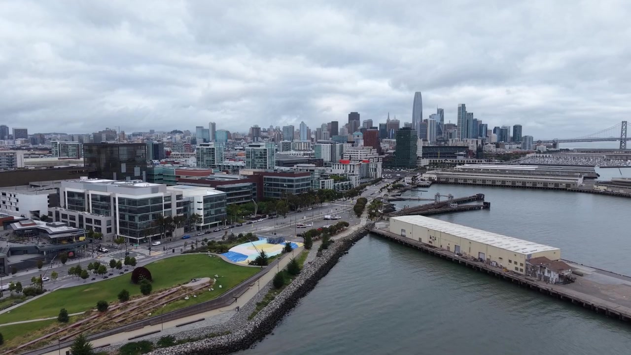 Una ripresa con drone in avvicinamento cattura l'iconico skyline di San Francisco, il lungomare e il Bay Bridge sotto cieli drammatici e coperti. Nitide riprese aeree rivelano l'architettura urbana e la vivace energia del porto