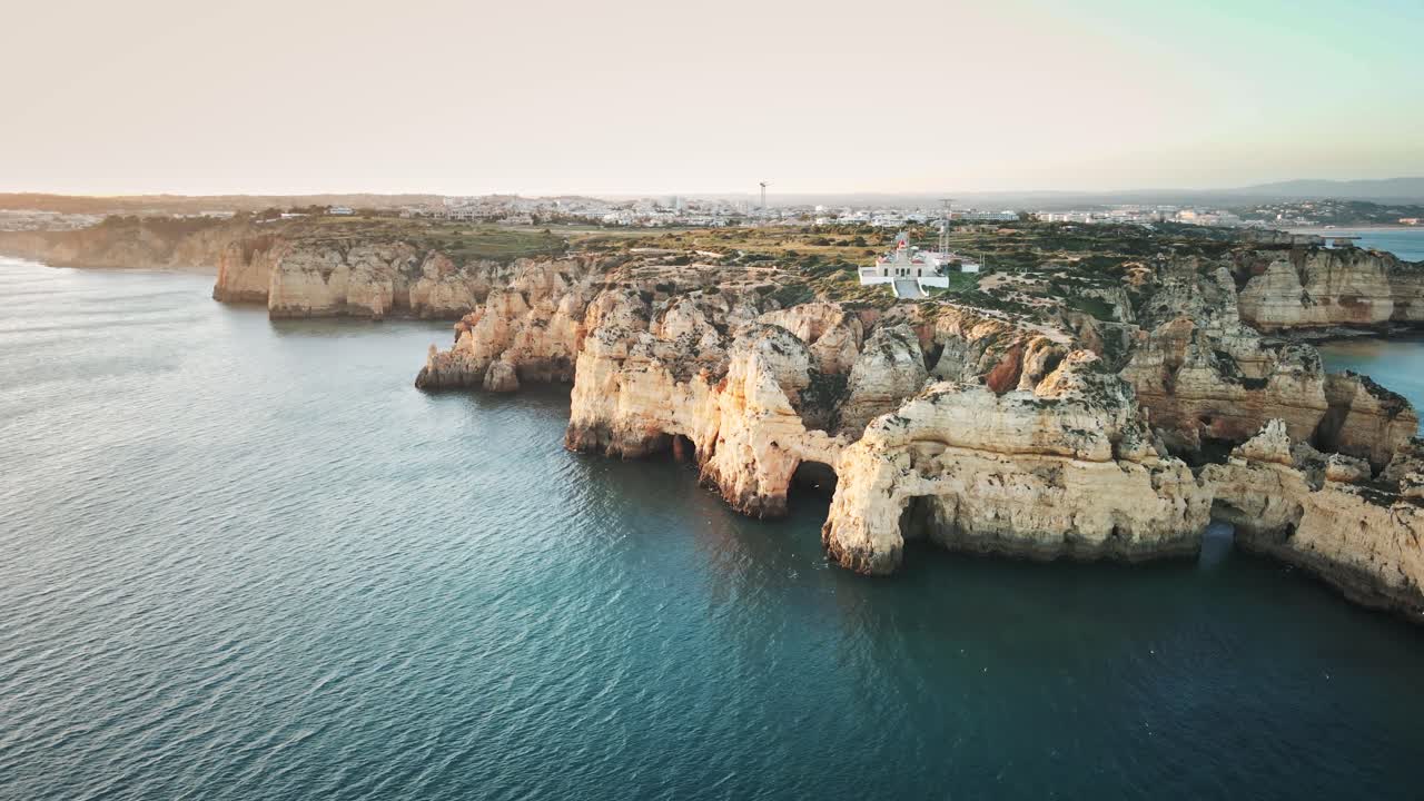 Camera spirals along Algarve’s cliffs and Ponta da Piedade lighthouse, unveiling sunlit rock formations, turquoise waters, scenic coastline, and distant town