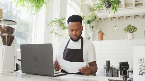 african american male cafe owner using laptop and looking at paperwork behind counter at cafe