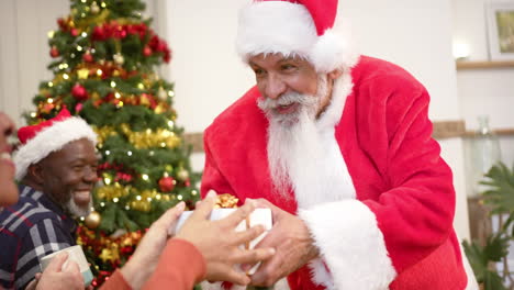 Fröhliche,-Abwechslungsreiche-Seniorenfreunde-Im-Weihnachtsmann-Outfit-Und-Weihnachtsmützen-Tauschen-Zu-Hause-Geschenke-Aus,-Zeitlupe