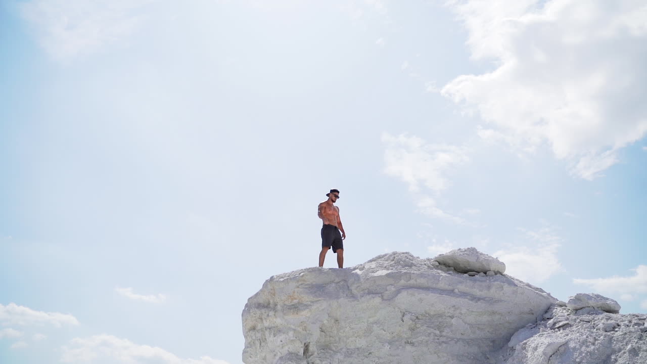Sportive man on the edge of the hill. Shirtless man in shorts under the light sky. Muscular athlete on the top of mount in summer. View from below. Slow motion.