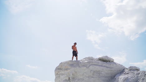 sportive man on the edge of the hill. shirtless man in shorts under the light sky. muscular athlete on the top of mount in summer. view from below. slow motion.