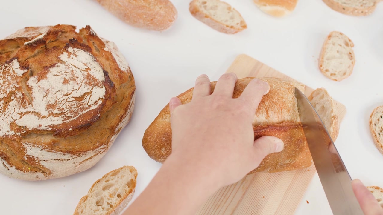 Top-down view of hands using a knife to slice crusty sourdough bread on white background.