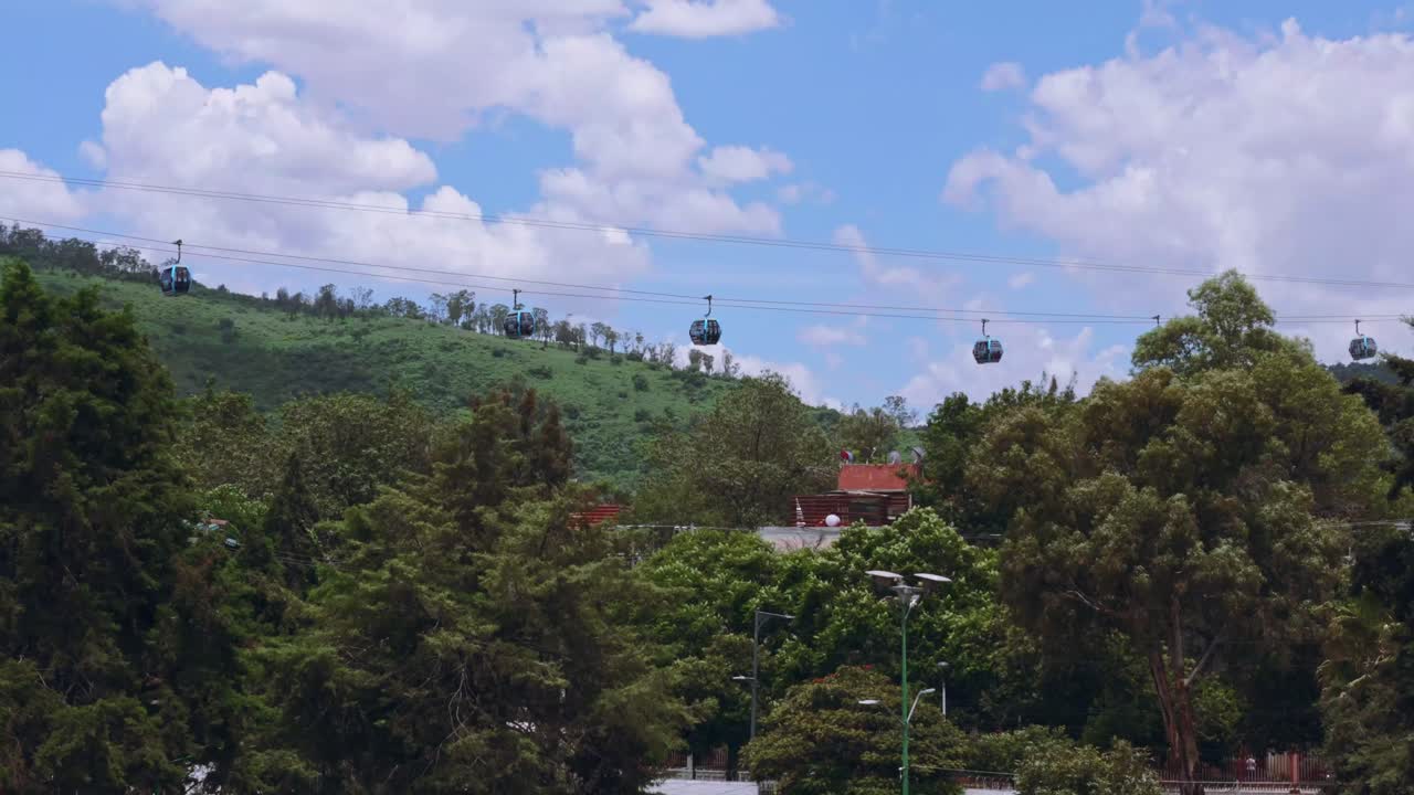 Cable cars over forested hillside under blue sky. Aerial zoom out reveals lush vegetation and urban rooftops in the distance.