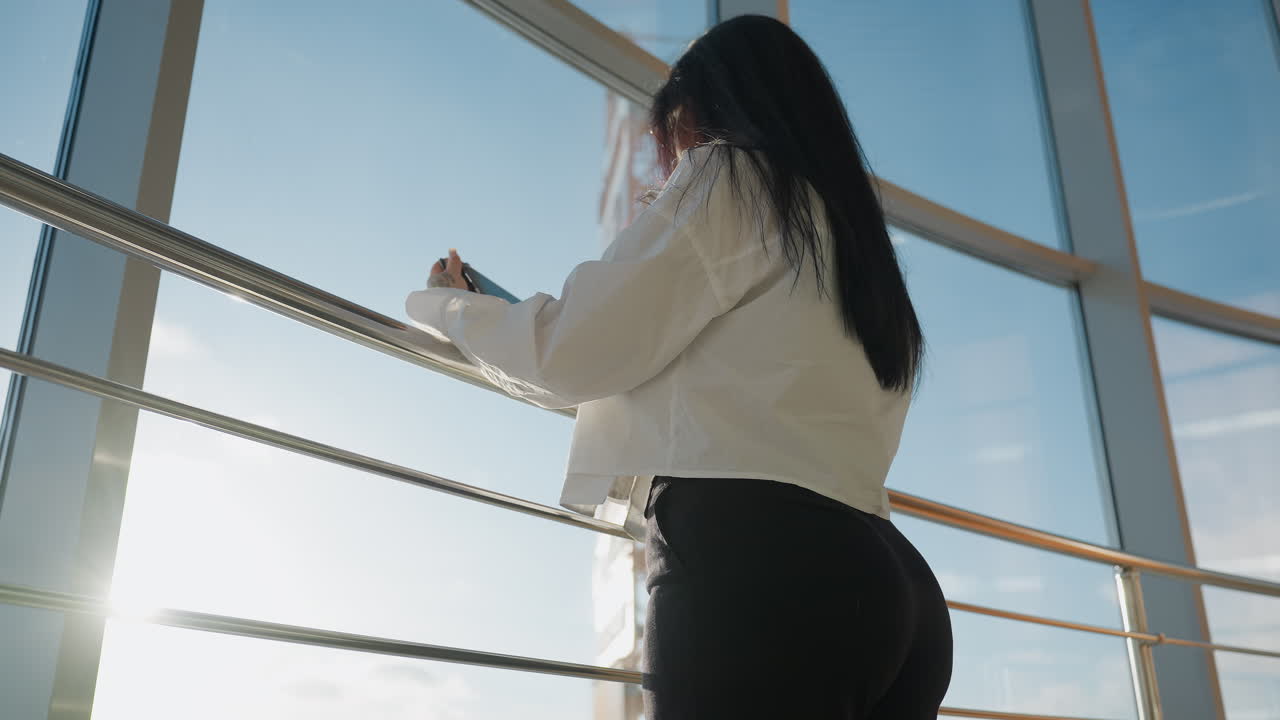Back view of woman in white shirt using tablet while standing by metal railing near large window, outdoor cityscape with urban structures visible through transparent glass under daylight